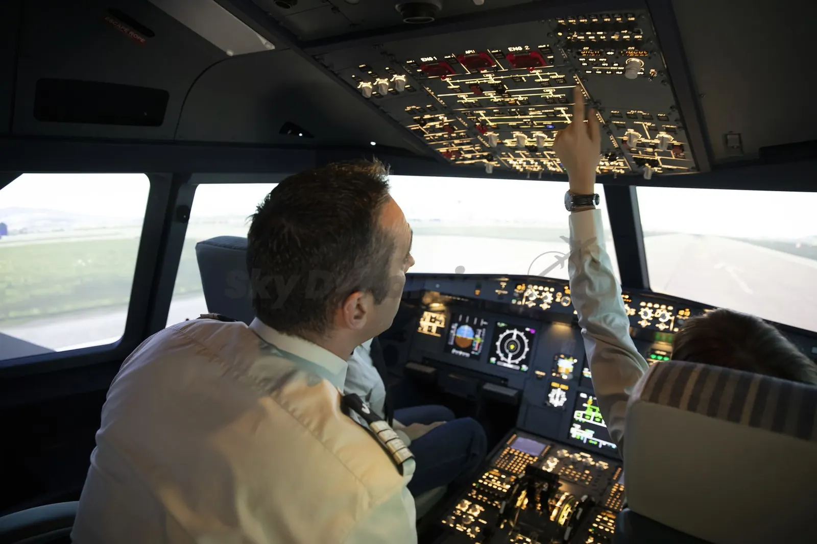 Two pilots on the flight deck of the SkyDynamics A320 FNPT II simulator, seen from behind, one reaching for the overhead panel